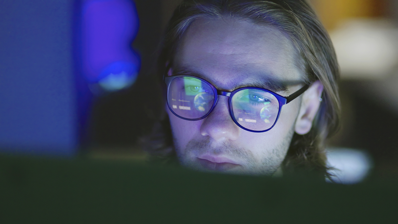 Close-up of a face of a young, long haired man, working at night in a dark office